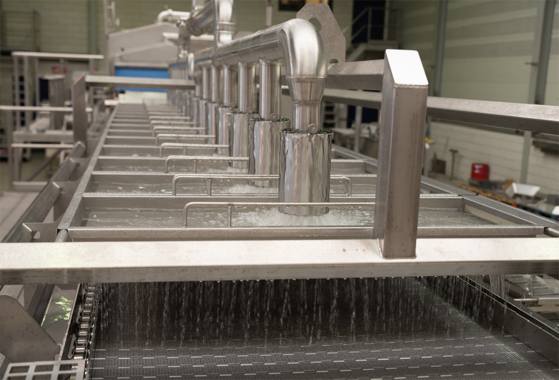 Close-up view of the OctoFrost IF Chiller system inside a food processing facility. Multiple stainless steel nozzles release a uniform rainshower of water over a conveyor belt below, illustrating hygienic and efficient cooling in action.